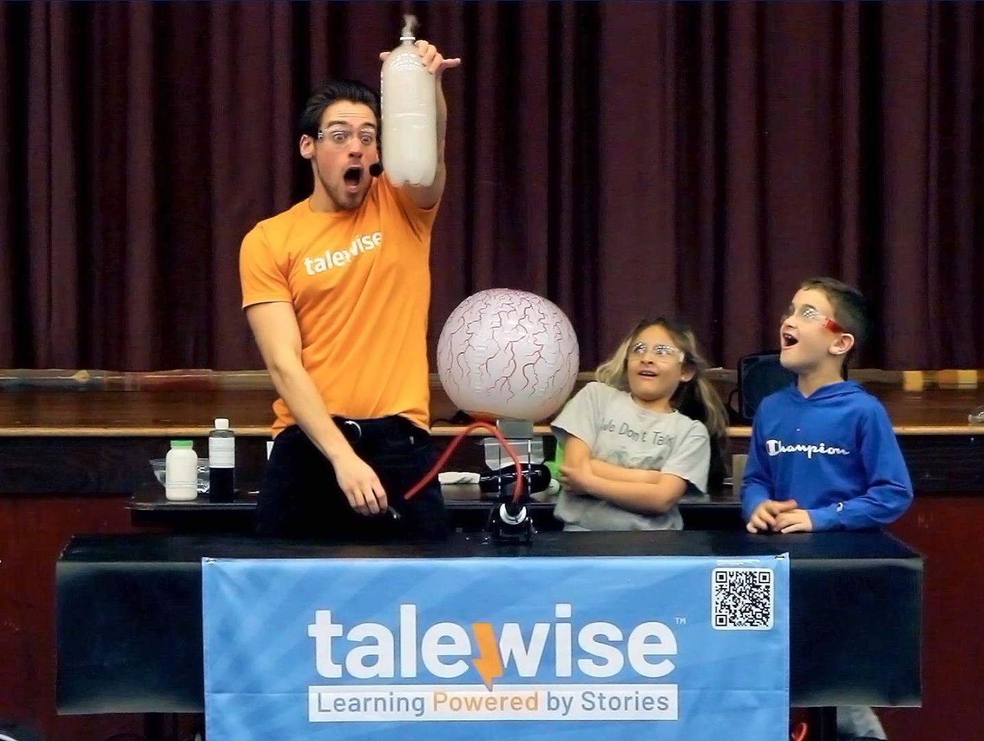 A performer from Talewise Science Heroes holds up a clear plastic bottle full of smoke with a shocked expression. Two kids watch with surprise and excitement.
